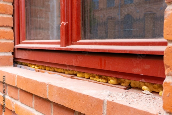 Fototapeta Installation of window with expanding foam insulation, red frame fitted into brick wall of building façade.