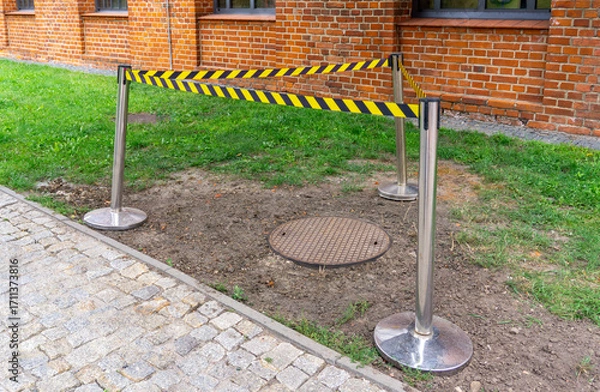 Fototapeta Safety barrier with warning tape securing a manhole cover on a lawn near cobblestone pavement and brick building wall.