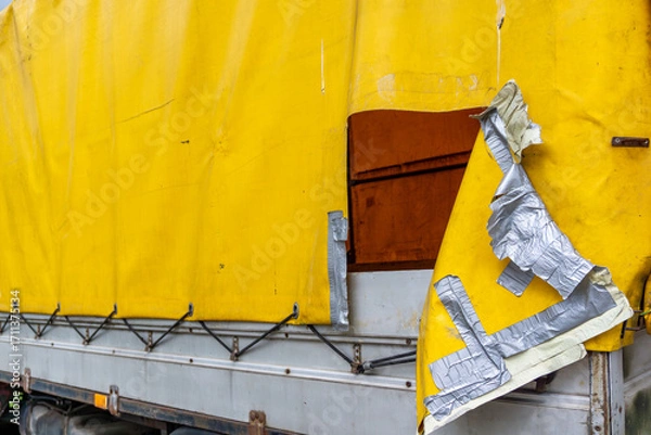 Fototapeta Damaged yellow truck tarpaulin, torn and patched with duct tape, visible metal trailer structure and supporting construction elements.