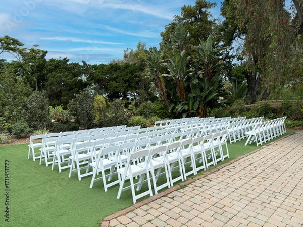 Fototapeta Empty wedding ceremony seating setup with white chairs on grass beside paved walkway in garden with large trees, greenery, and open sky