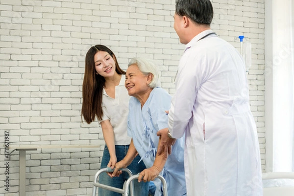 Fototapeta Smiling senior patient using a walker receives support from a doctor and family caregiver in a bright clinic, symbolizing rehabilitation, mobility assistance, and compassionate healthcare.