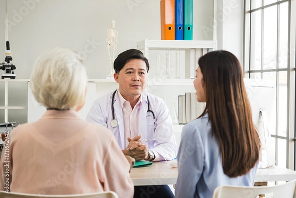 Fototapeta Doctor speaking with two patients during medical consultation in a bright clinic office, discussing healthcare and treatment options across a desk, stethoscope visible.