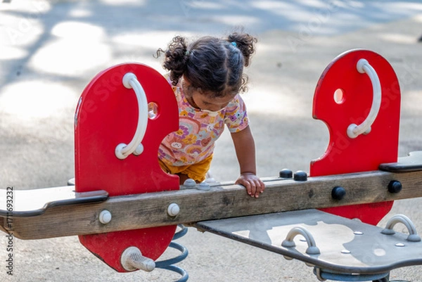 Fototapeta African American girl playing on a colorful playground seesaw, showcasing childhood joy and innocence, surrounded by a sunny outdoor environment with playful energy