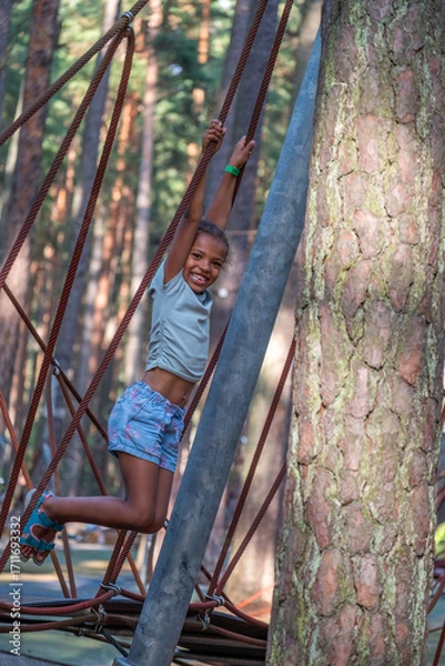 Fototapeta African American girl joyfully swinging on ropes in a forest playground, surrounded by tall trees, capturing the essence of childhood happiness and playful innocence