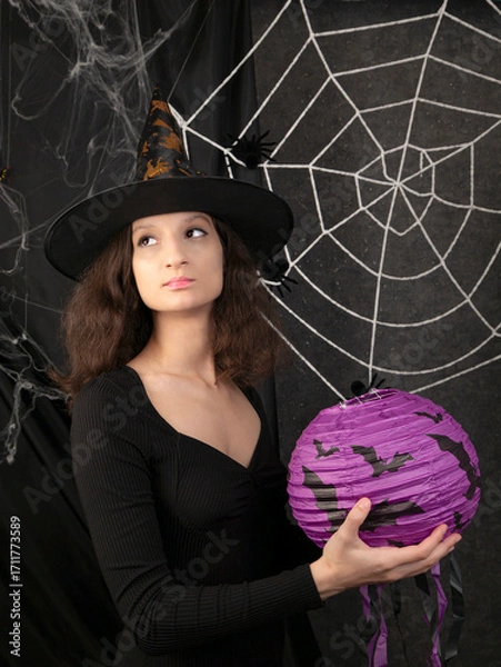 Obraz Beautiful witch girl in a witch's cap holds a lilac lantern in her hands against the background of a white web on Halloween