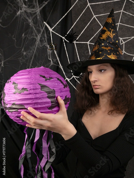 Obraz Beautiful witch girl in a witch's cap holds a lilac lantern in her hands against the background of a white web on Halloween