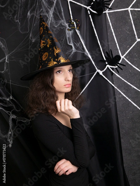 Obraz Beautiful girl witch in a witch's cap thoughtfully against the background of a web on Halloween