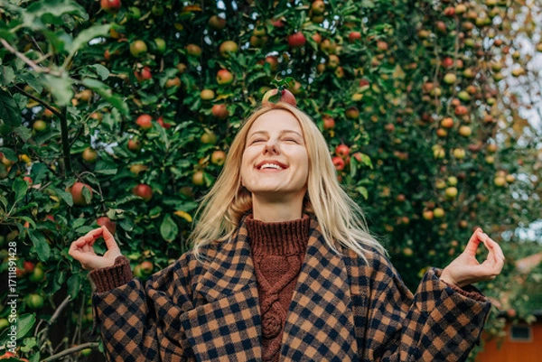 Fototapeta European woman stands by an apple tree in an autumn orchard, playfully balancing a ripe red apple on her head