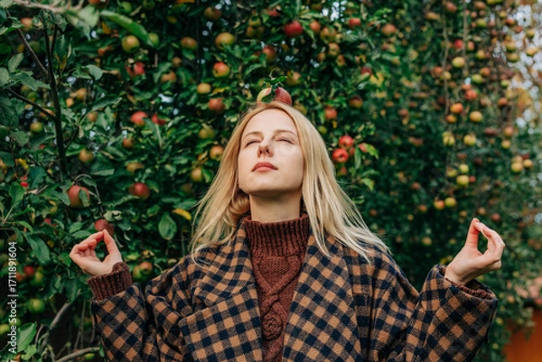 Fototapeta European woman stands by an apple tree in an autumn orchard, playfully balancing a ripe red apple on her head