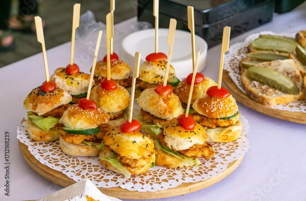 Fototapeta Plate with mini burgers on skewers topped with cherry tomatoes, next to sandwiches with pickles on wooden tray.