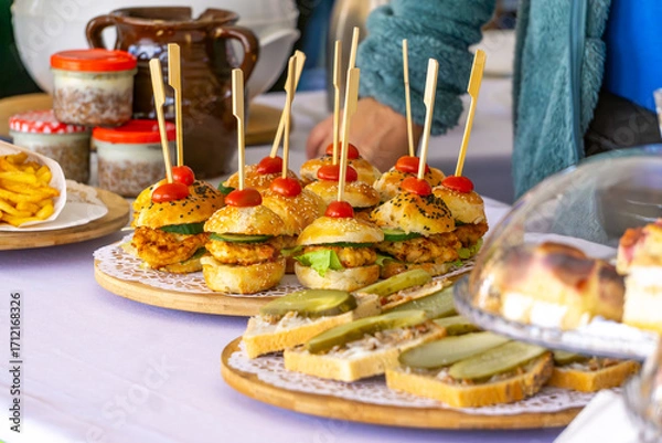 Fototapeta Table with appetizers and snacks, mini burgers, sandwiches with pickles, fries, and food jars visible in background.