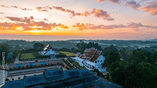 Fototapeta Aerial view of a traditional temple surrounded by greenery under a vibrant sunrise sky. The golden and blue hues of twilight highlight the intricate architecture and peaceful ambiance of the scene