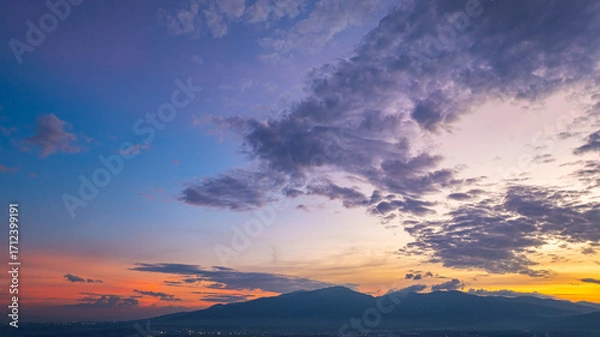 Fototapeta A breathtaking aerial panoramic view of a vibrant sunset illuminating the sky above a lush green hilltop with a small structure, surrounded by scattered clouds and distant landscapes.