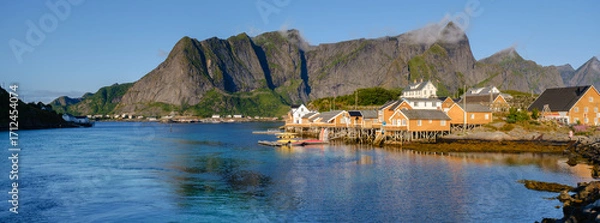 Fototapeta Bright colors of historic wooden cabins line the peaceful waterfront of Sakrisoy in Lofoten, Norway. Majestic mountain peaks rise dramatically under a clear blue sky, creating a serene atmosphere.