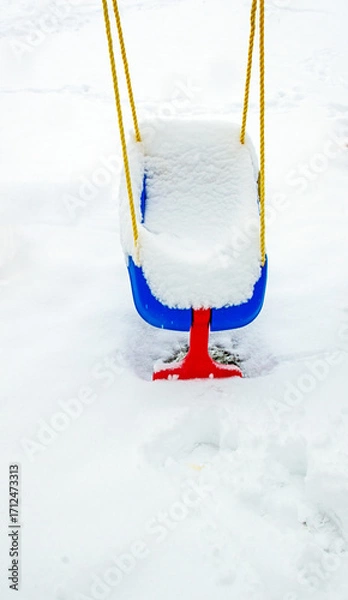 Fototapeta White fluffy snow fills a blue and red baby swing. The yellow ropes add more eye catching color to the winter scene.