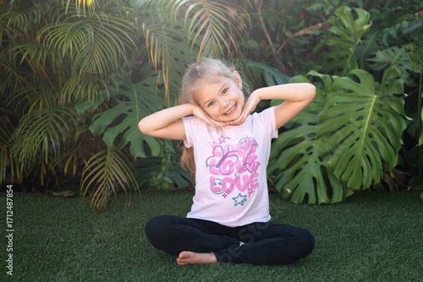 Fototapeta Happy little girl smiling and posing playfully with arms under her chin while sitting in front of lush green tropical leaves, expressing carefree childhood energy and authentic joy.