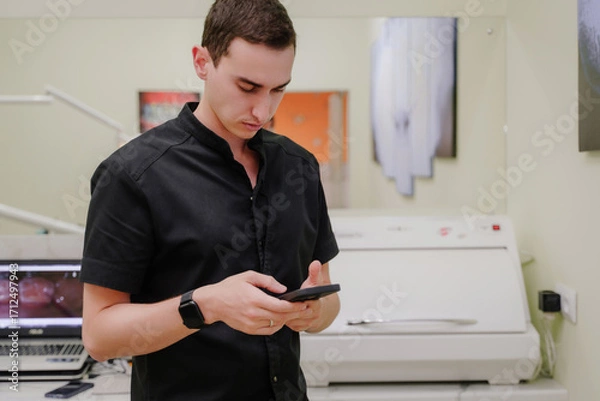 Obraz The dentist conducts correspondence on a smartphone. The doctor is standing in the office and looking at something on the phone. A serious doctor in a black uniform.