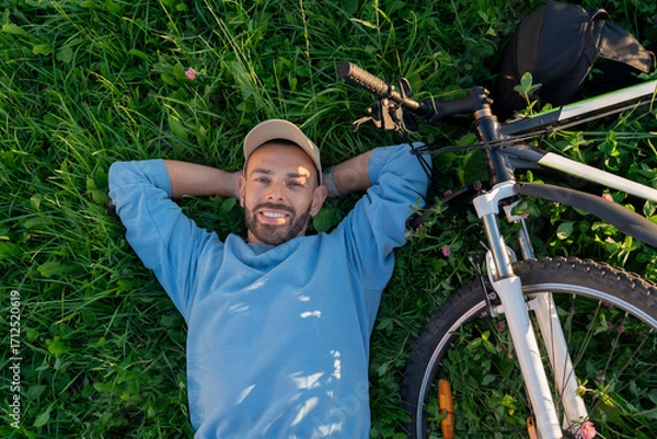 Fototapeta A smiling man lies on his back in the grass near his bicycle. He is resting with his hands behind his head on a sunny day