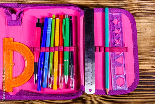 Fototapeta Different school stationeries (pens, pencils, ruler and protractor) in a pink pencil box. Top view