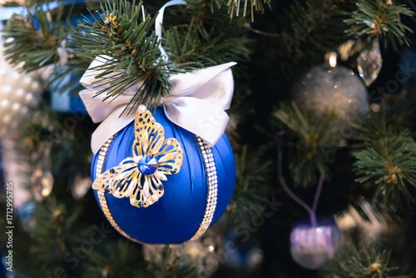 Fototapeta Elegant blue Christmas ball decorated with a shiny butterfly and white satin bow, hanging on a festive tree with sparkling lights and needles