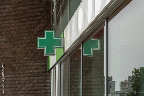 Fototapeta green cross, a common symbol for a pharmacy, is mounted on the corner of a brick building. The sign is reflected in the large glass windows of the modern facade