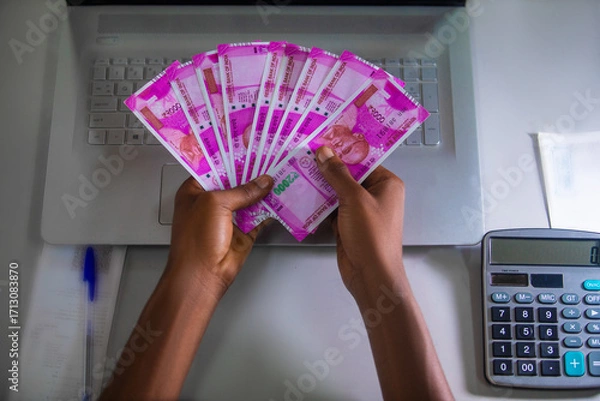 Fototapeta Dark skinned African person sitting holding Indian Rupee notes with calculator, laptop and pen on table desk