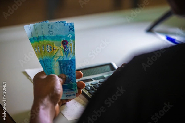 Fototapeta Dark skinned African person in black top working on desk holding Kuwaiti Dinar notes in hand and pressing punching calculator with receipt on table. accounting concept