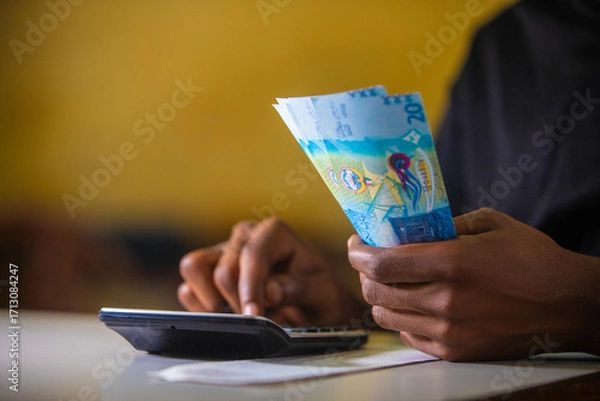 Fototapeta Dark skinned African person in black top working on desk holding Kuwaiti Dinar notes in hand and pressing punching calculator with receipt on table. accounting concept. 