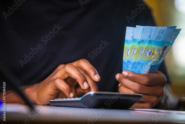 Fototapeta Dark skinned African person in black top working on desk holding Kuwaiti Dinar notes in hand and pressing punching calculator with receipt on table.  accounting concept