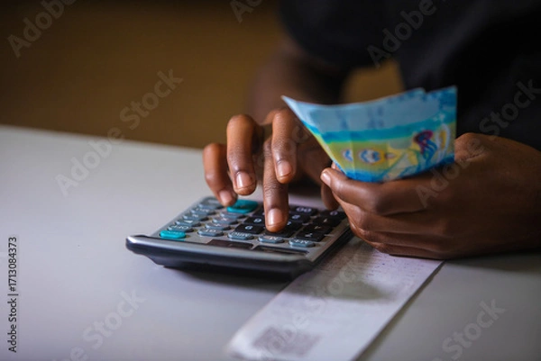 Fototapeta Dark skinned African person in black top working on desk holding Kuwaiti Dinar notes in hand and pressing punching calculator with receipt on table.  accounting concept