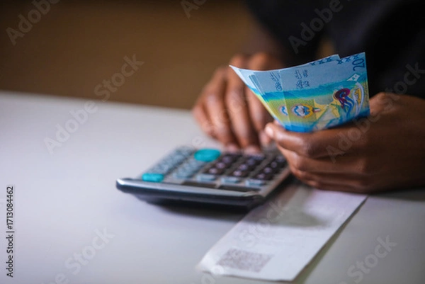 Fototapeta Dark skinned African person in black top working on desk holding Kuwaiti Dinar notes in hand and pressing punching calculator with receipt on table. accounting concept.