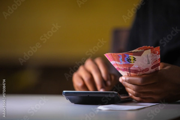 Fototapeta Dark skinned African person working on white desk pressing punching calculator and holding Ghanaian cedi notes in hand. Accounting concept