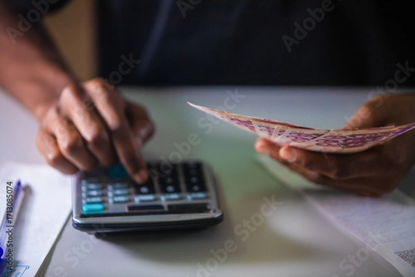 Fototapeta Top view of Dark skinned African person working on white desk pressing punching calculator and holding Ghanaian cedi notes in hand with more money on desk. accounting concept