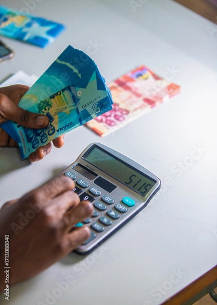 Fototapeta Dark skinned African person working on desk holding Ghanaian cedi notes in hand and pressing punching calculator with some notes on table. Top view accounting concept