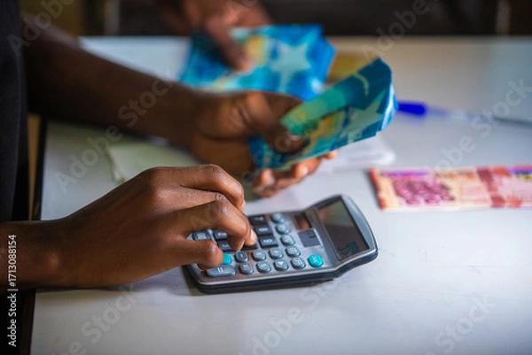 Fototapeta Dark skinned African person working on desk holding Ghanaian cedi notes in hand and pressing punching calculator with some notes on table. Top view accounting concept