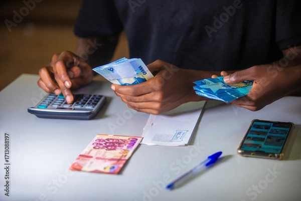 Fototapeta Dark skinned African person working on desk writing in notebook and holding Ghanaian cedi notes in hand with some notes on table. Top view accounting concept
