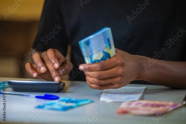 Fototapeta vertical shot of Dark skinned African person working on desk holding counting Ghanaian cedi notes money with some notes on table. accounting concept. 