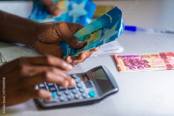 Fototapeta Dark skinned African person working on desk holding Ghanaian cedi notes in hand and pressing punching calculator with some notes on table. Top view accounting concept