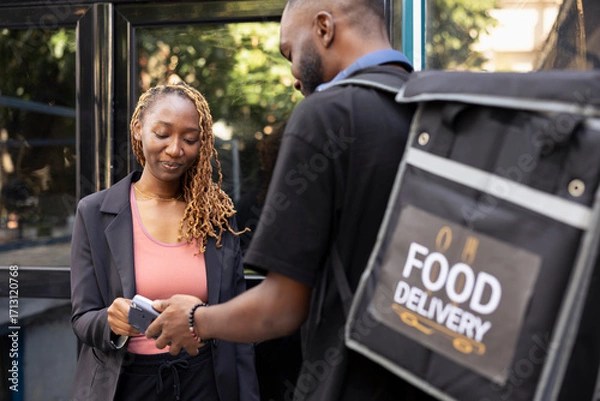 Fototapeta Black client completes a fast food purchase with contactless payment at the doorstep, courier holds pos terminal steady as the electronic transaction is processed. Wireless purchase.