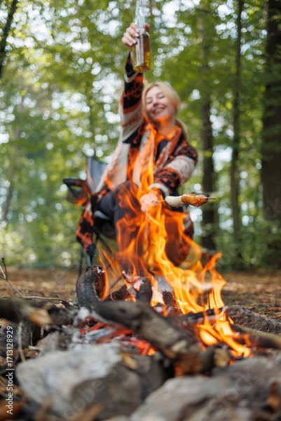 Fototapeta Happy woman raising beer and roasting sausage over campfire flames outdoors