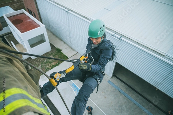 Fototapeta Female firefighter rescuer beginning the descent of a huge wall using the rappelling device in a multi-story building.