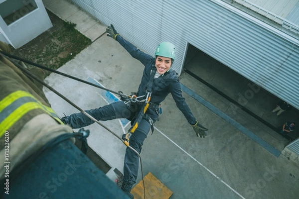 Fototapeta Female firefighter rescuer descending a huge wall with the rappelling device in a multi-story building, hanging without hands and smiling at the camera.