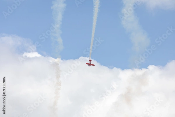 Fototapeta A restored World War II era red biplane airplane performing aerobatic maneuvers in the sky during an air show event, leaving a trail of smoke against a cloudy background in Hillsboro, Oregon, USA
