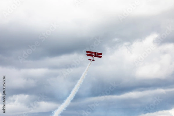 Fototapeta A restored World War II era red biplane airplane performing aerobatic maneuvers in the sky during an air show event, leaving a trail of smoke against a cloudy background in Hillsboro, Oregon, USA