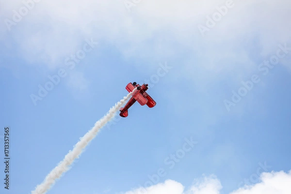Fototapeta A restored World War II era red biplane airplane performing aerobatic maneuvers in the sky during an air show event, leaving a trail of smoke against a cloudy background in Hillsboro, Oregon, USA