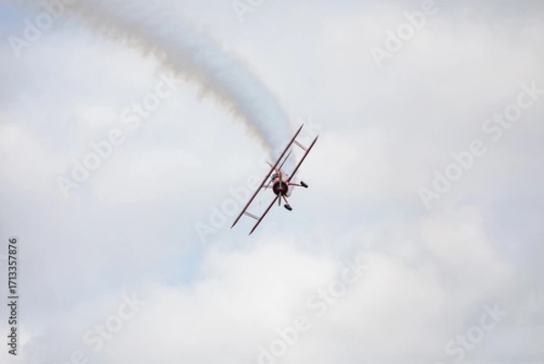Fototapeta A restored World War II era red biplane airplane performing aerobatic maneuvers in the sky during an air show event, leaving a trail of smoke against a cloudy background in Hillsboro, Oregon, USA