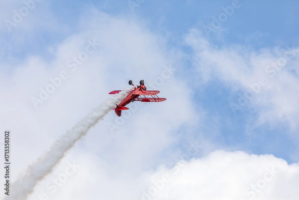 Fototapeta A restored World War II era red biplane airplane performing aerobatic maneuvers in the sky during an air show event, leaving a trail of smoke against a cloudy background in Hillsboro, Oregon, USA