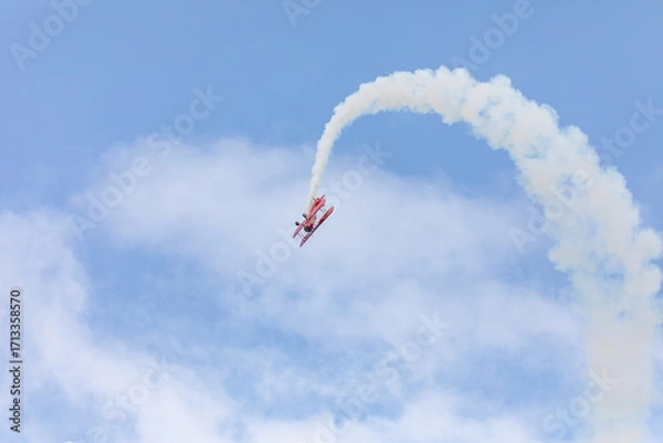 Fototapeta A restored World War II era red biplane airplane performing aerobatic maneuvers in the sky during an air show event, leaving a trail of smoke against a cloudy background in Hillsboro, Oregon, USA