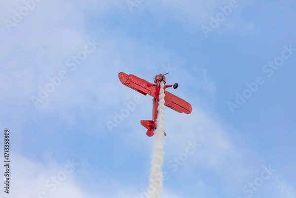 Fototapeta A restored World War II era red biplane airplane performing aerobatic maneuvers in the sky during an air show event, leaving a trail of smoke against a cloudy background in Hillsboro, Oregon, USA
