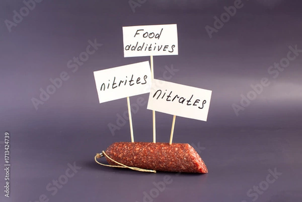 Fototapeta A fragment of smoked sausage in a package and the warning labels "nitrites" and "nitrates" in close-up on a dark background